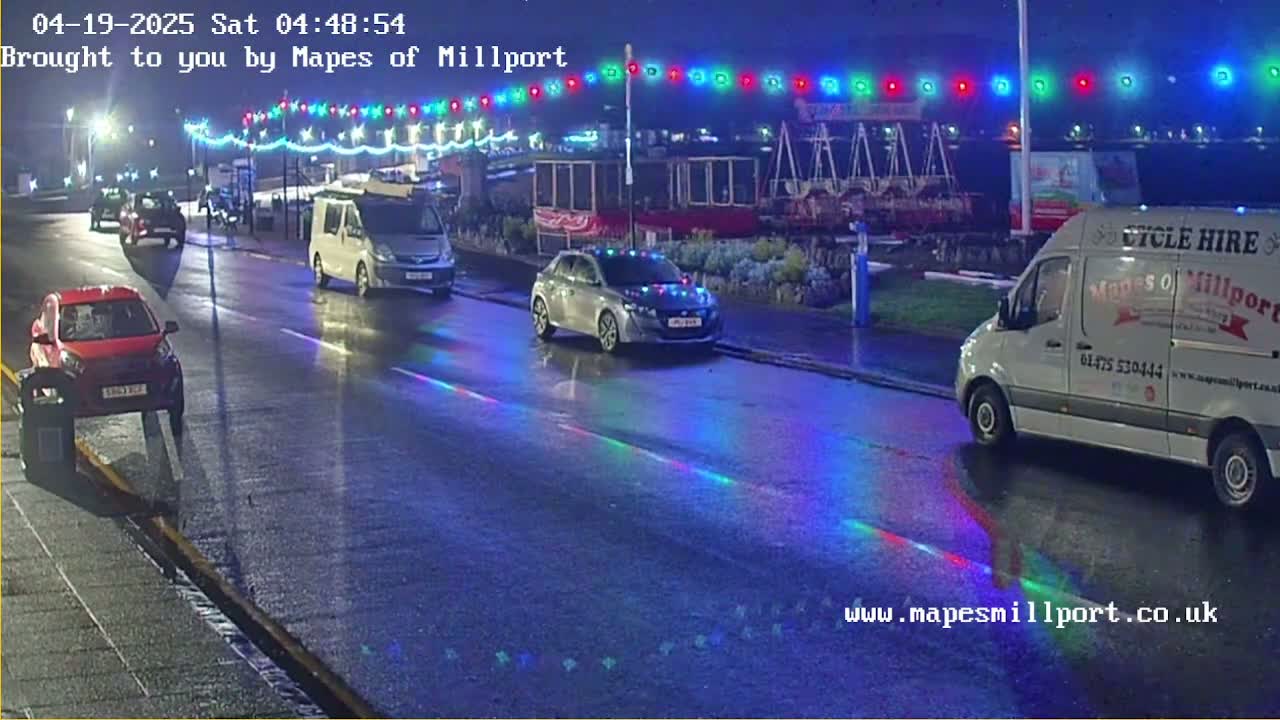 At night, several vehicles are parked and driving on a wet road under a string of multicolored lights.