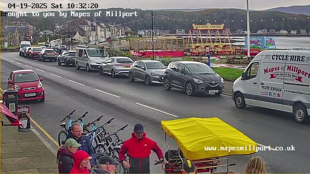 A street scene shows several cars parked along the side of a road, with people standing near bicycles and a yellow-covered cart, under overcast skies.