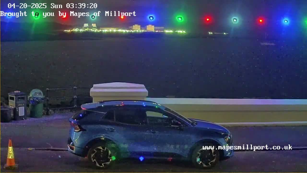 A blue SUV with decorative lights is parked near a low wall overlooking a dark body of water, with city lights and colored lights in the distance under a nighttime, possibly snowy sky.