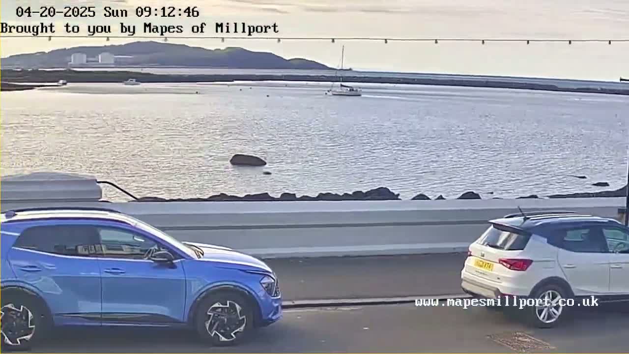 A blue and a white SUV are parked near a calm body of water under a partly cloudy sky.