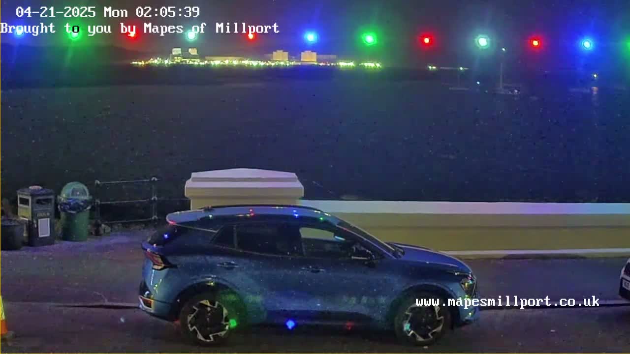 A blue SUV is parked at night near a waterfront, with lights visible in the distance and light rain or snow falling.