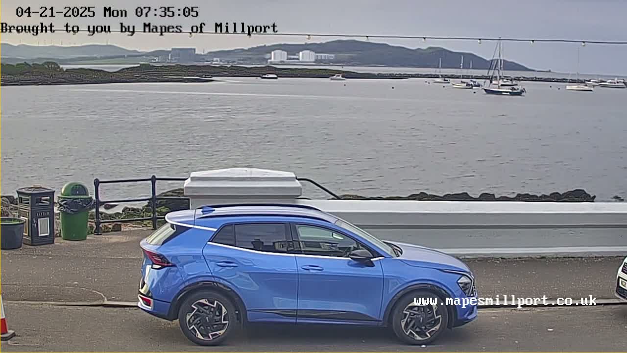 A blue SUV is parked near a waterfront, overlooking a calm body of water with several boats and a distant shoreline under an overcast sky.