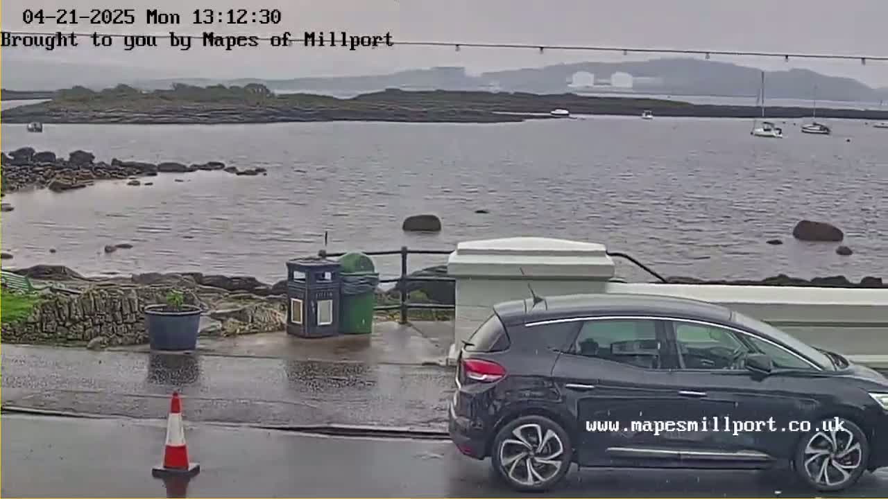 A dark-colored car is parked near a rocky shoreline overlooking calm, gray water under an overcast sky.