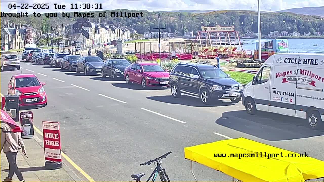 Cars are parked along a street next to a seaside town on a sunny day, with a carousel and a van visible in the background.