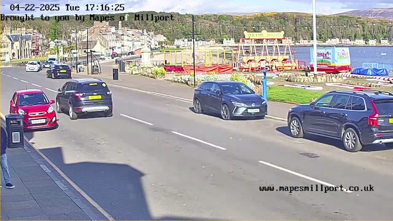 Several cars drive down a seaside road on a sunny day, past a playground with a carousel.