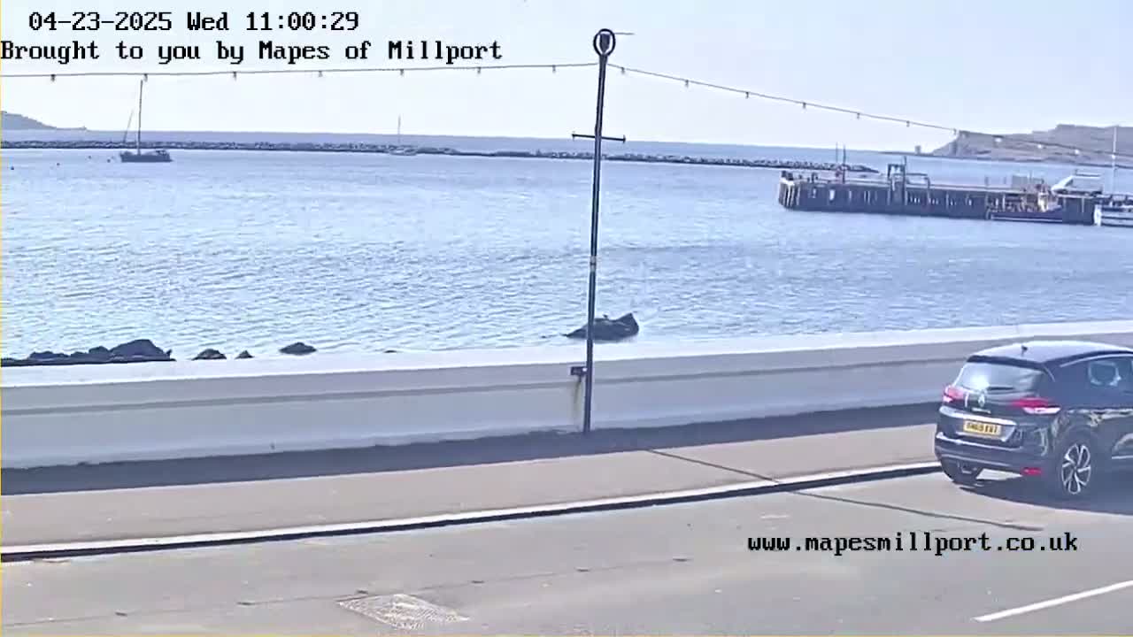 A dark-colored SUV is parked near a waterfront wall overlooking a calm body of water on a sunny day, with a sailboat and a dock visible in the distance.