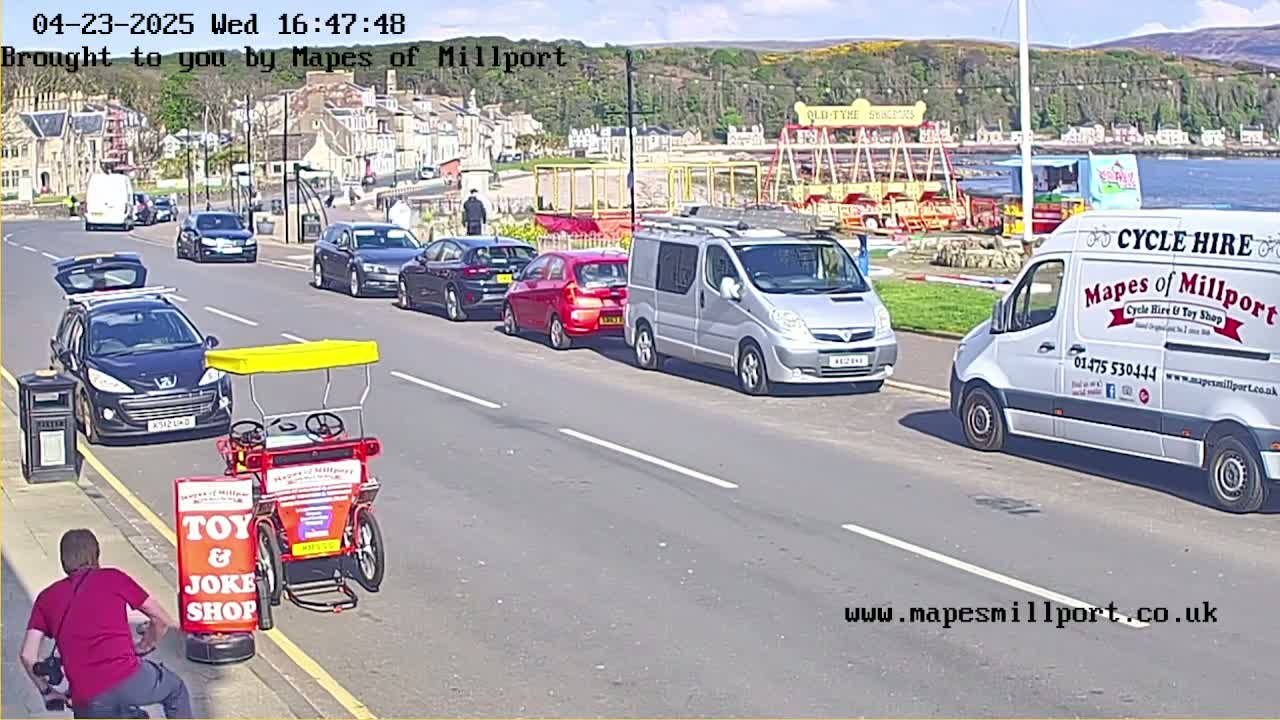 A sunny seaside street scene shows several cars parked along the curb, with a red toy and joke shop surrey bike in the foreground, and a person crouching near it.