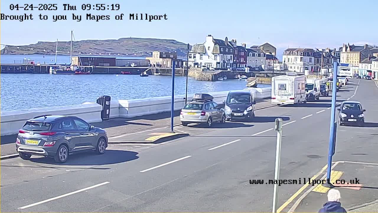 A sunny day shows several vehicles driving along a seaside road with a harbor and buildings in the background.