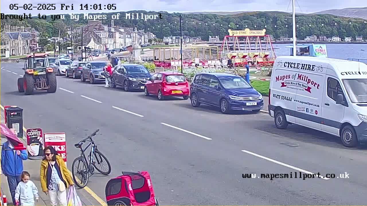 A tractor, several cars, a van, and a bicycle are parked along a street near a seaside town on a sunny day.