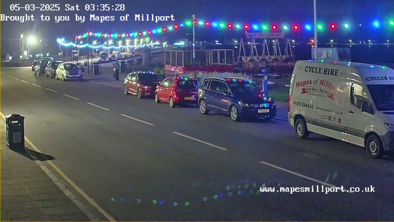 At night, several cars are parked along a road under a string of multicolored lights, with a white van parked on the right side of the road.