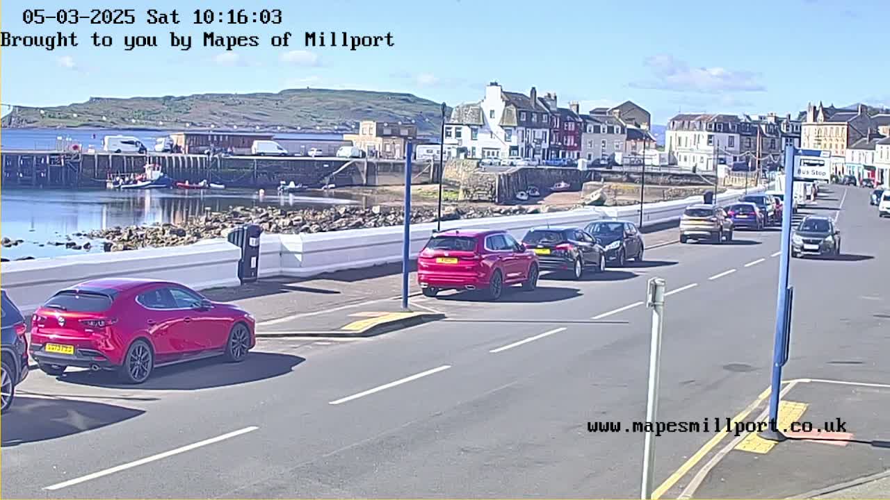 Several cars are parked and driving along a seaside road on a sunny day, with a harbor and buildings in the background.