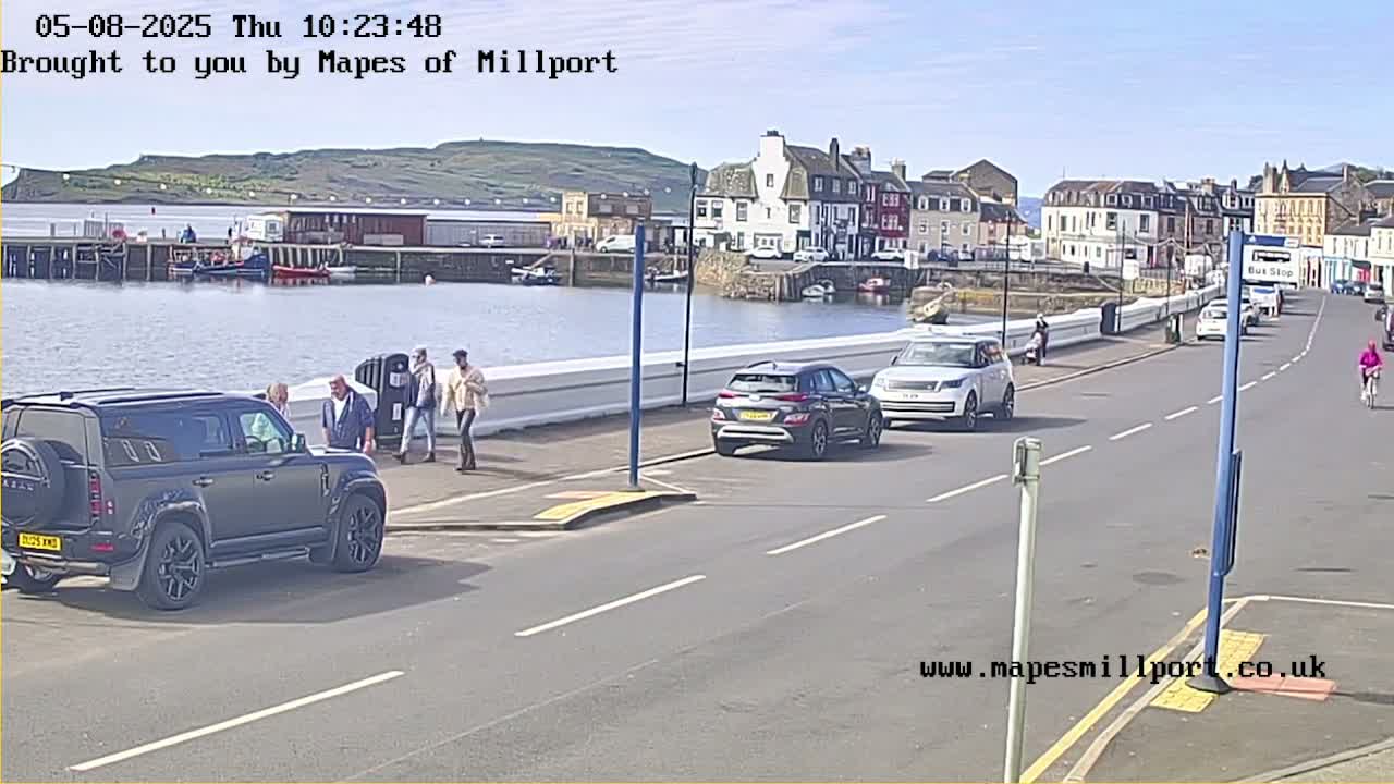 A sunny day shows a harbor with several cars parked along a road near buildings, and a few pedestrians walking by.