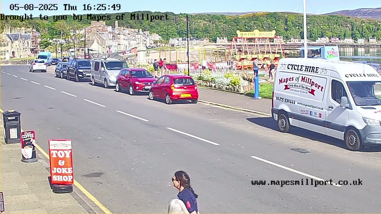 A sunny seaside street scene shows several cars parked along the roadside near a carousel and a white van, with a woman walking along the sidewalk.