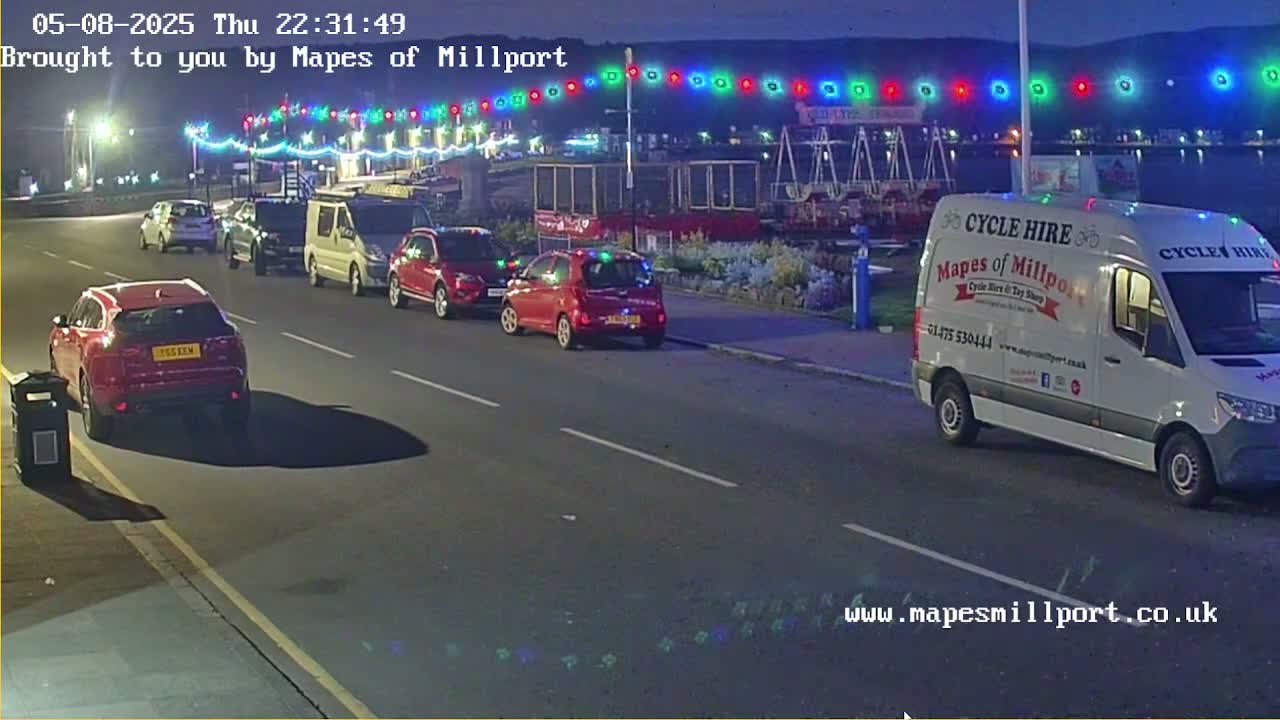 Several cars and a van are parked along a street at night under a string of colorful lights.