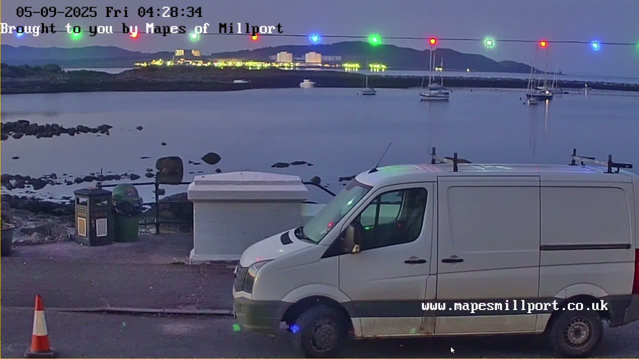 A white van is parked near a rocky shore at dusk, overlooking a calm body of water with several boats and a distant town lit up.
