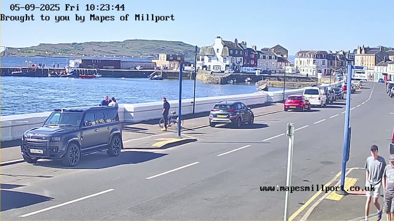 A sunny waterfront scene shows several cars parked along a road next to a harbor with buildings and people visible in the background.