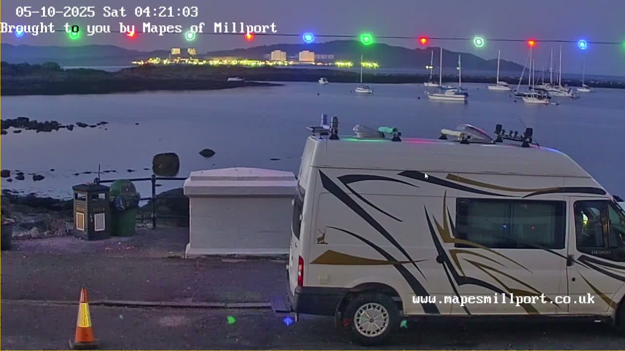 A white van with gold-and-black striping is parked near a body of water at twilight, with several sailboats visible in the distance under a clear, dark sky.