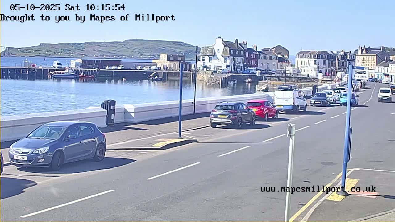 Several cars are parked along a road next to a harbor on a sunny day.