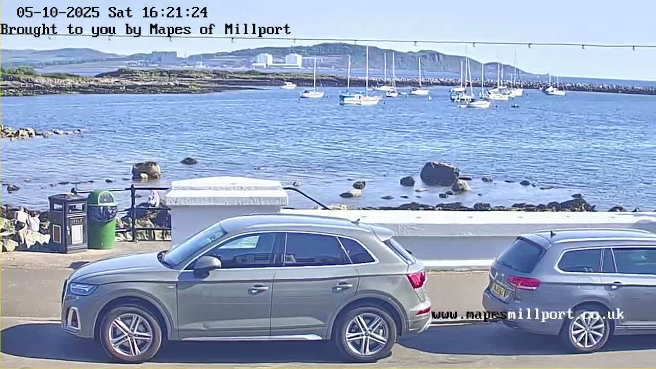 Two cars are parked near a body of water on a sunny day, with several sailboats visible in the distance and a rocky shoreline.