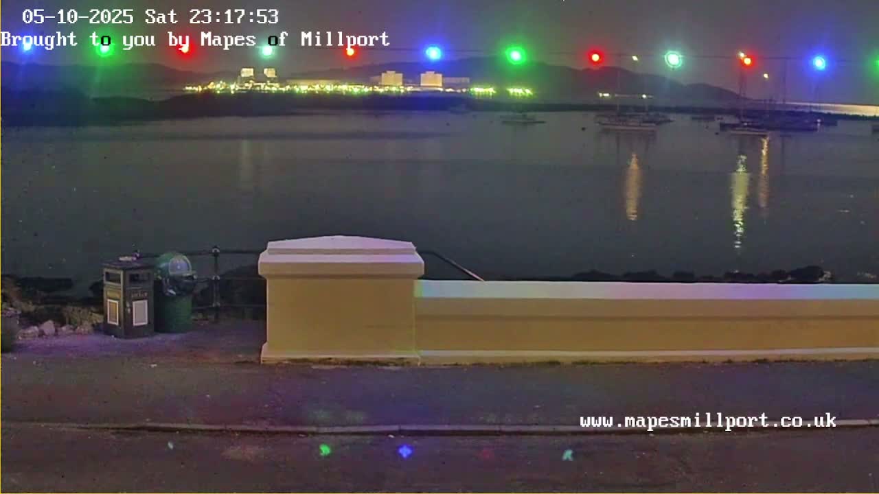 At night, a calm body of water reflects the lights of a distant town and several boats, all viewed from behind a low wall.