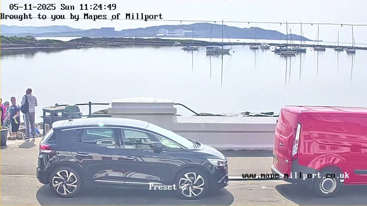 A sunny day shows a calm body of water with several sailboats moored, a black SUV, and a red van parked near a waterfront area where a few people stand.