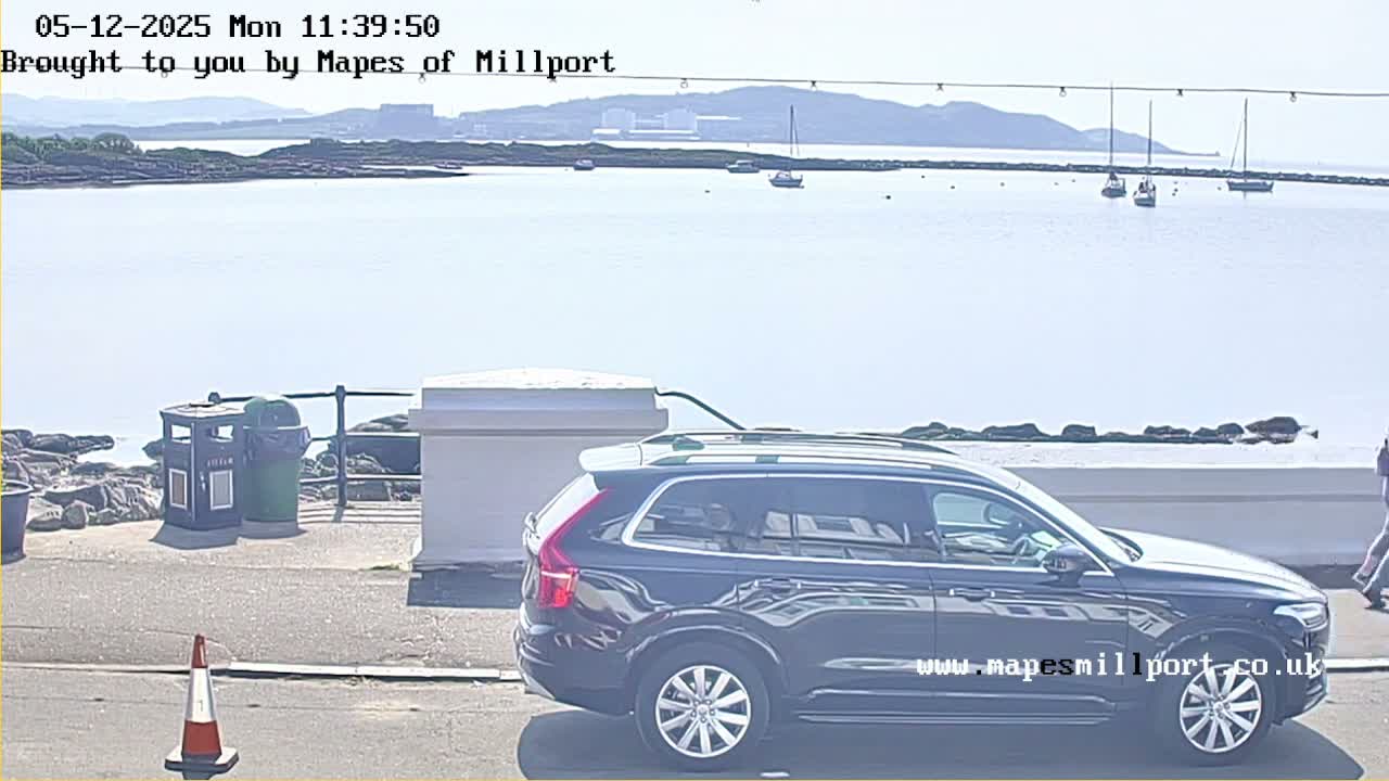 A dark-colored SUV is parked near a rocky shoreline on a bright, sunny day with several sailboats visible in the calm water.