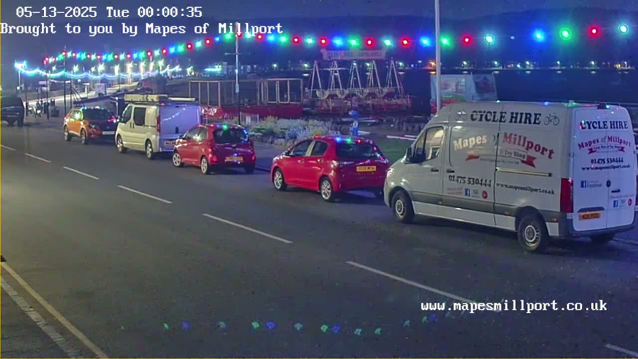 Several cars and vans are parked on a street at night under a string of multicolored lights.