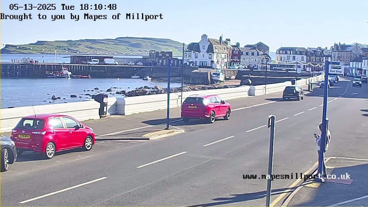 A sunny seaside town with cars parked along a road, a harbor with boats, and a person standing on the sidewalk.