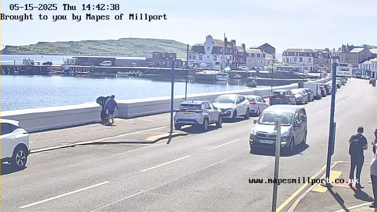 A sunny seaside town scene shows parked cars lining a road next to a harbor with several boats, and a few pedestrians walking near a low wall.