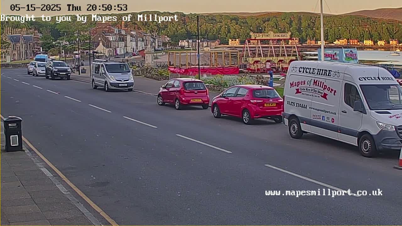 Several cars and a van are driving on a road in front of a seaside town at dusk, with hills in the background.