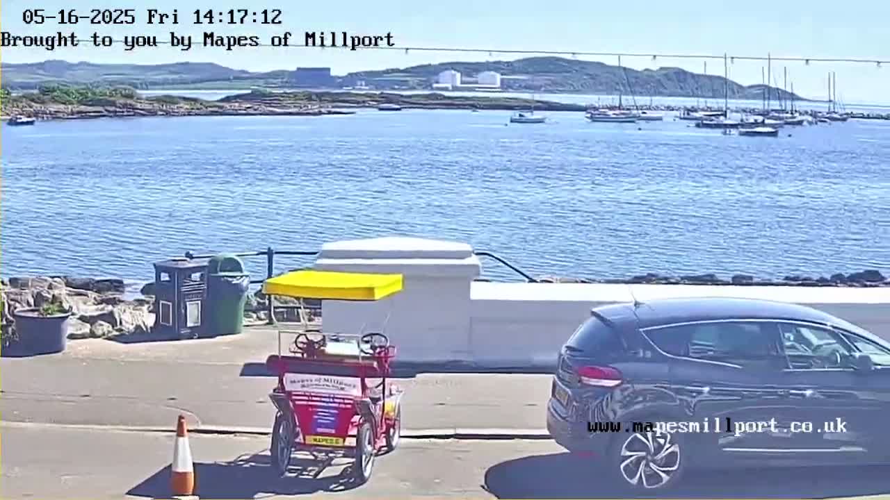 A sunny waterfront scene shows a calm body of water with boats in the distance, a parked car, and a surrey bike near a seawall.