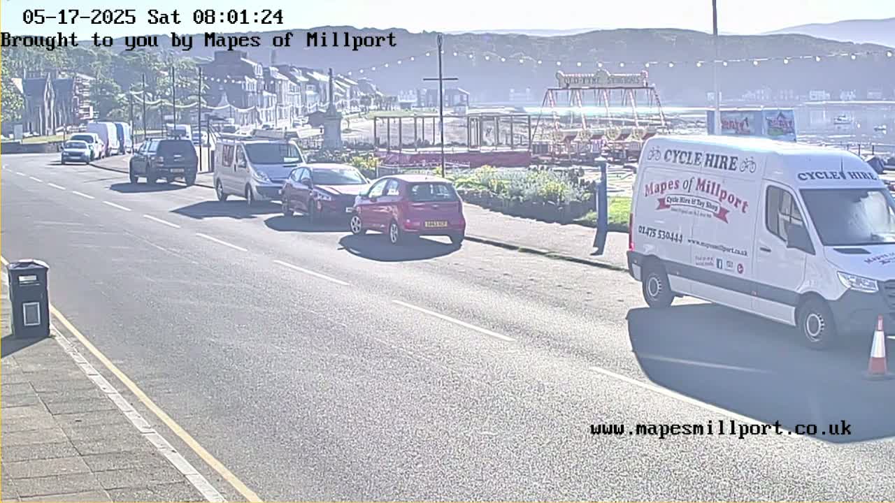 A sunny seaside street scene shows several cars and vans driving past a small amusement park.
