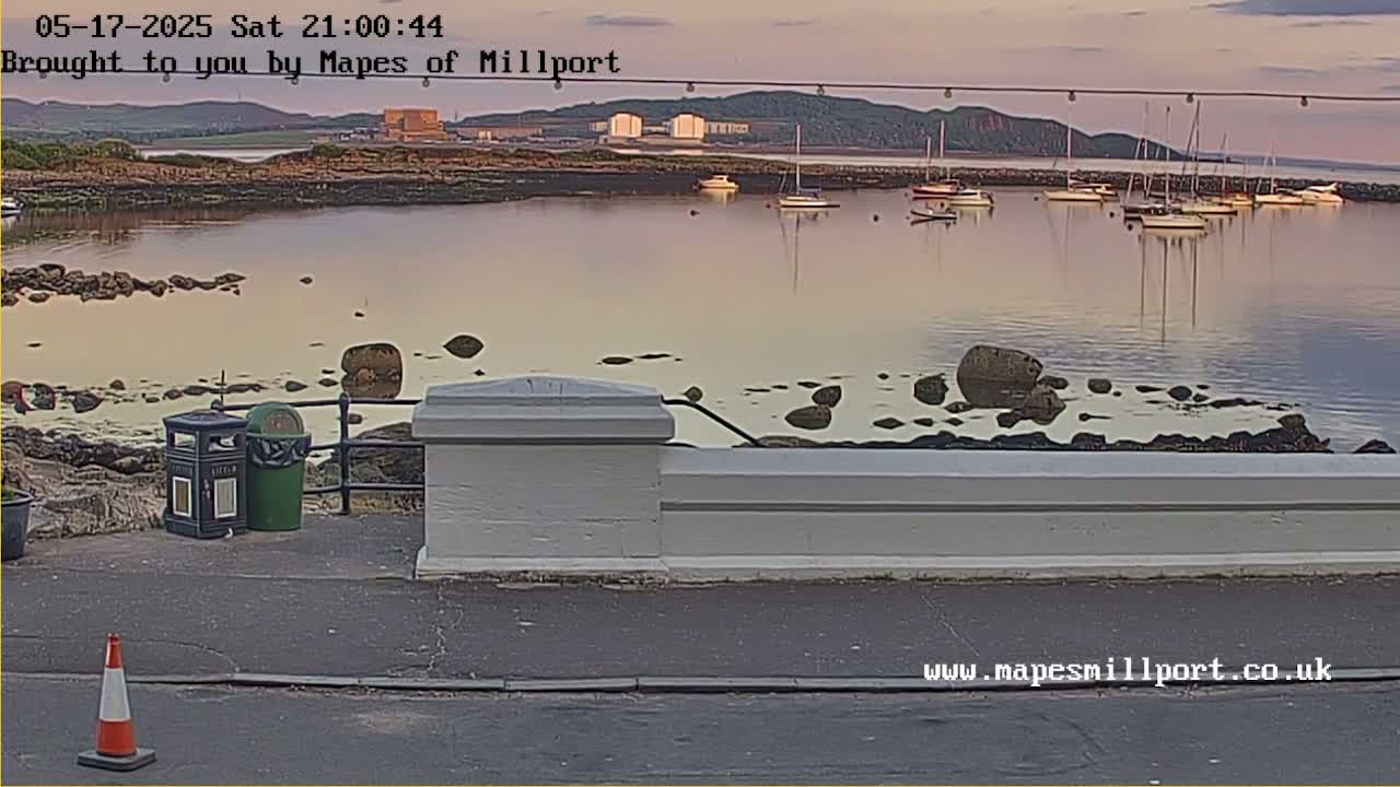 A calm harbor at dusk, with several sailboats moored near a rocky shore and a low white wall.