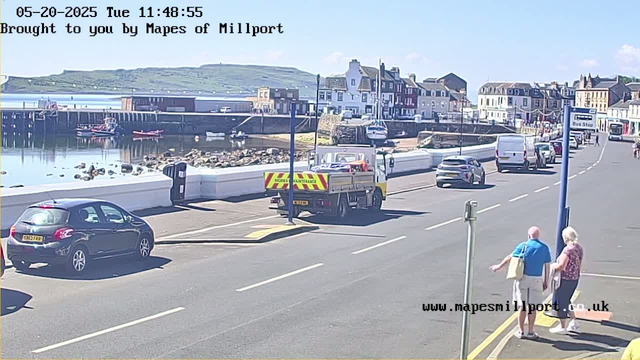 A sunny day shows a harbor with boats, a roadway with several vehicles including a maintenance truck, and two people standing near a bus stop in front of buildings.