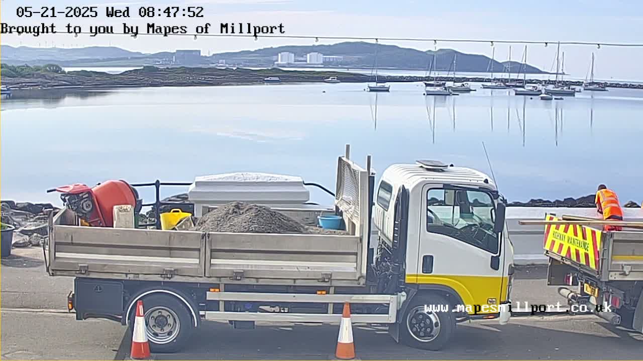 A white flatbed truck carrying construction materials is parked near a body of water with several sailboats in the background under a mostly clear sky.