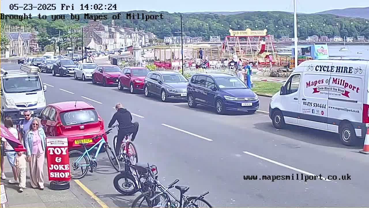 A sunny seaside street scene shows several parked cars, a few pedestrians, a man riding a bicycle, and a cycle hire van parked on the side of the road, near a carousel.