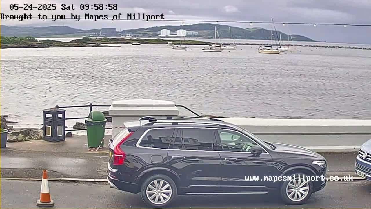 A dark-colored SUV is parked near a waterfront with several sailboats in the distance under an overcast sky.