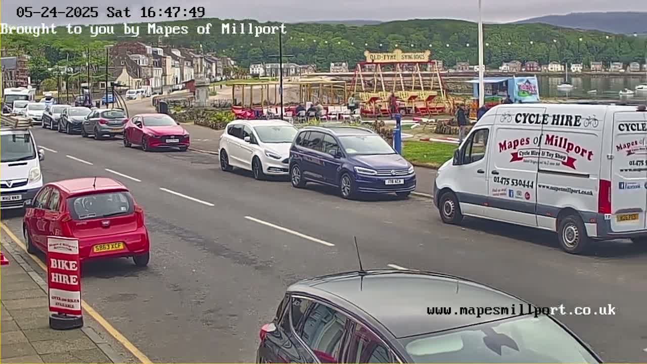 Several cars are parked along a street near a waterfront, with a white van and a carousel visible in the background under an overcast sky.