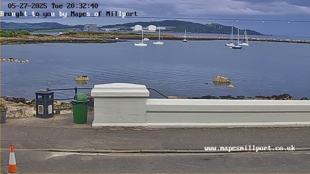 A calm bay with several sailboats is viewed from behind a low white wall on an overcast day.