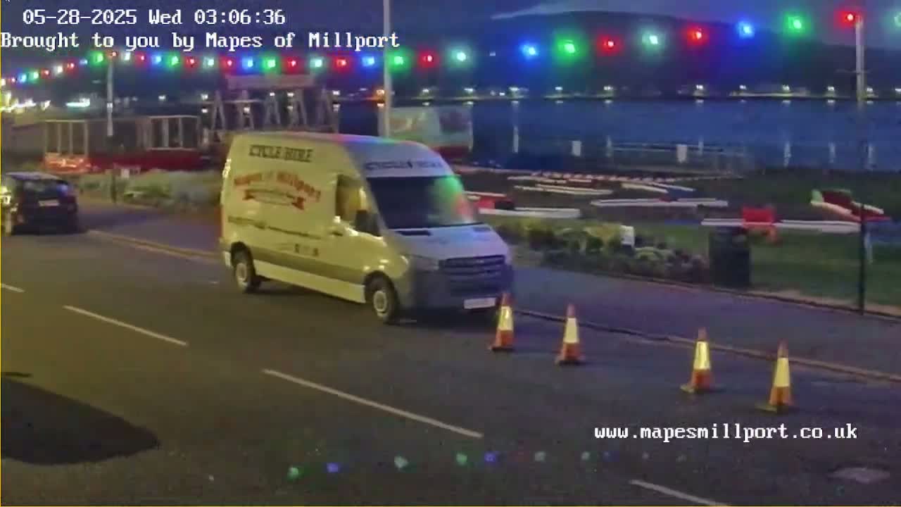 A light-colored van is parked on a dark street at night, under a string of colorful lights, with traffic cones nearby.