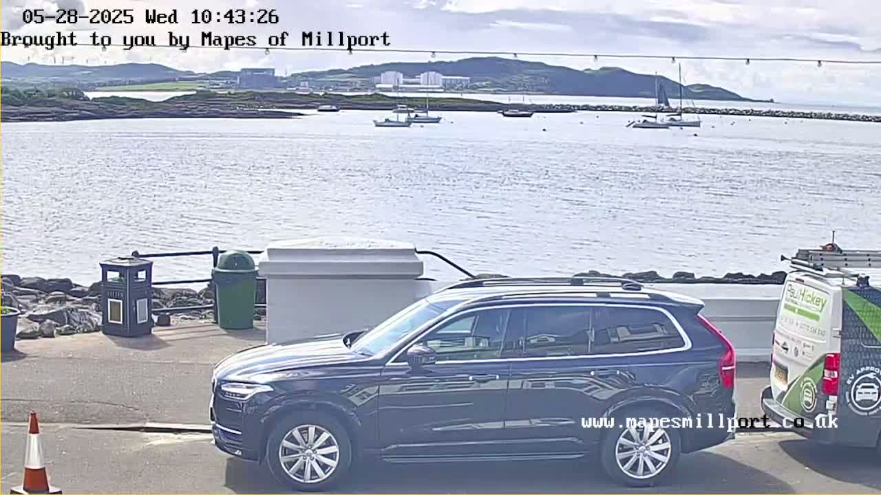 A dark-colored SUV is parked next to a van near a body of water on a partly sunny day, with several boats visible in the distance.