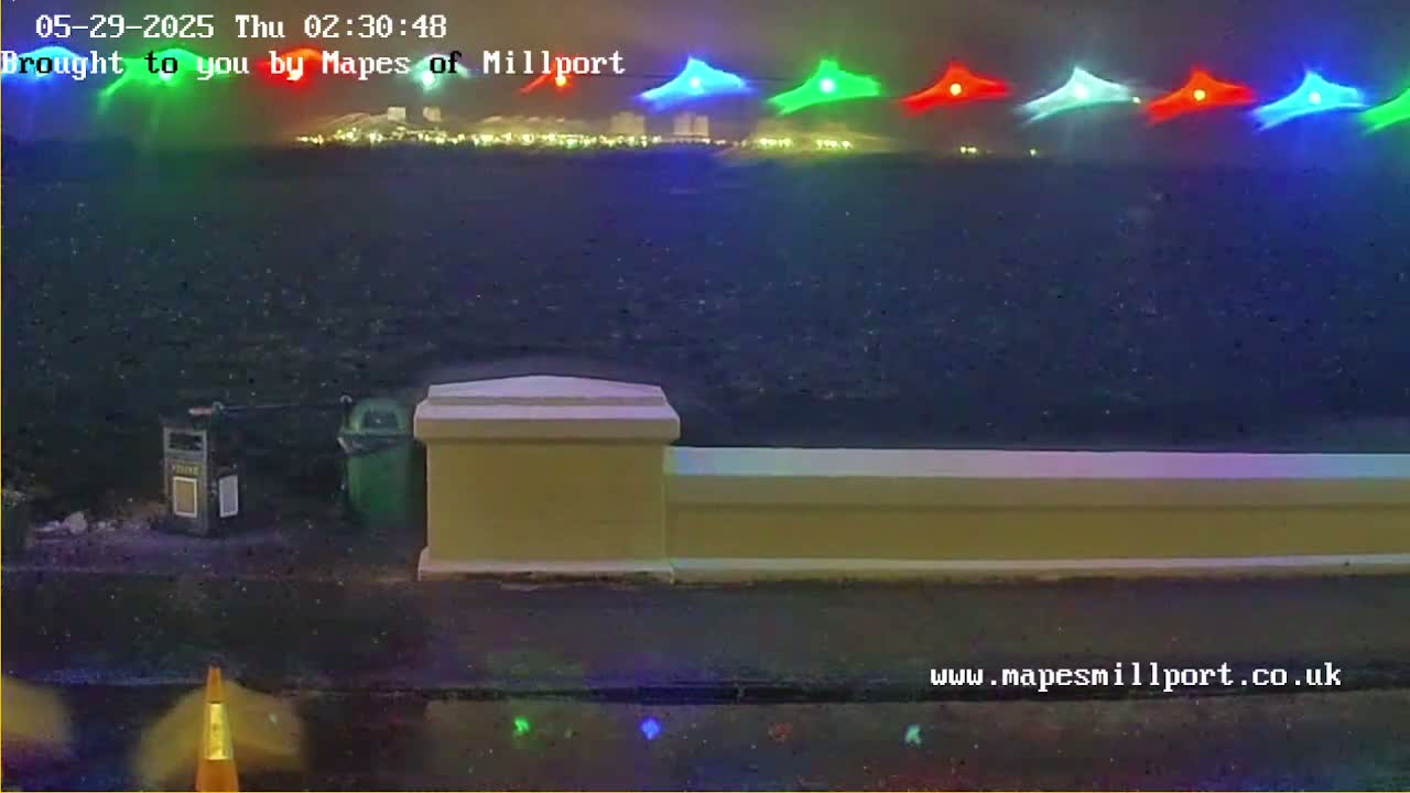A nighttime, wet scene shows a low wall beside a body of water with colorful lights in the distance.