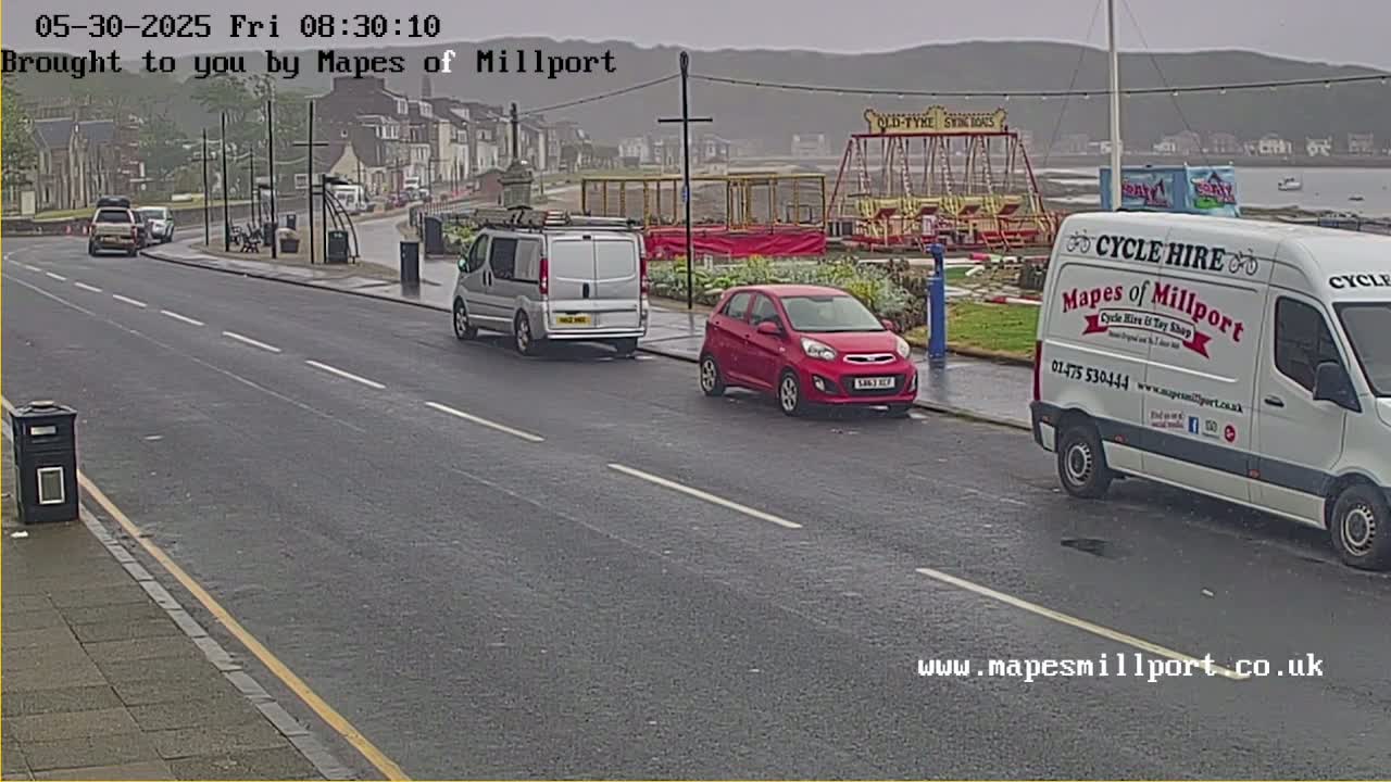 A wet, overcast day shows a seaside road with several parked cars, a van, and a small carousel in the background.