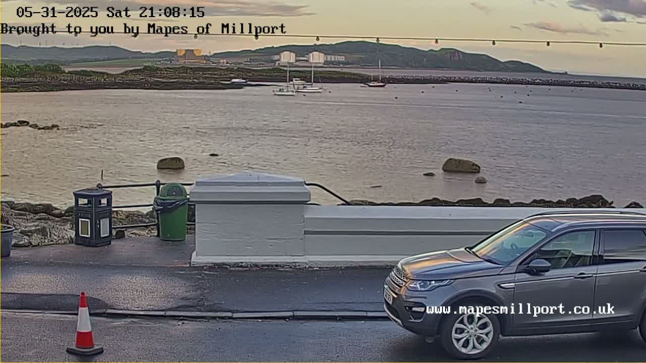 A gray SUV is parked near a low wall overlooking calm water where several boats are moored under a partly cloudy evening sky.