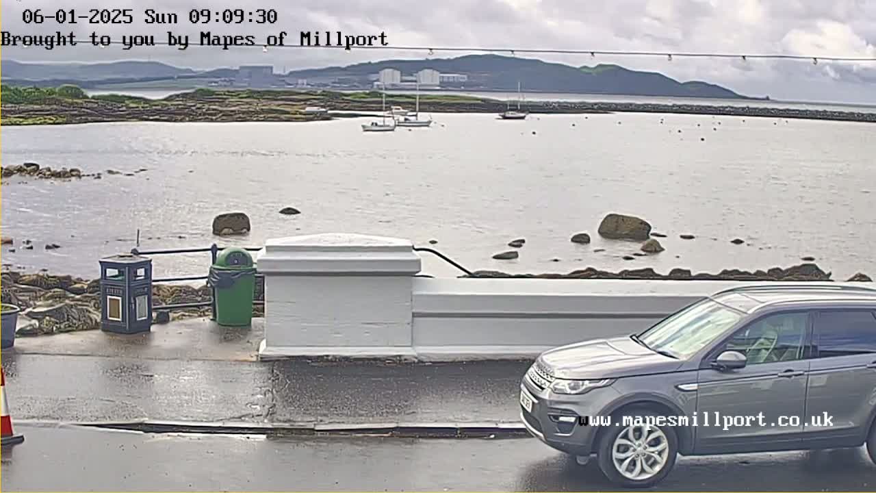 A gray SUV is parked near a low white wall overlooking a calm body of water with several boats and rocky shoreline under an overcast sky.