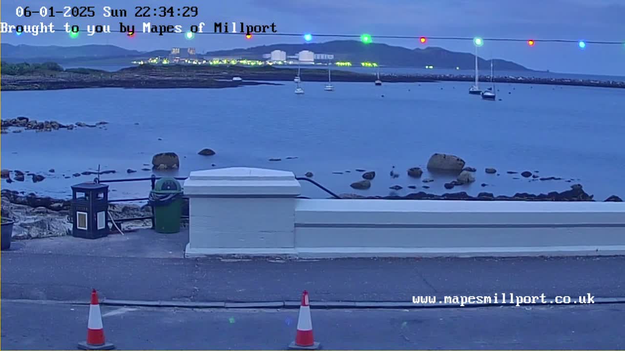 A twilight view of a calm body of water with several boats, rocks near the shore, and a low white wall with traffic cones in the foreground.