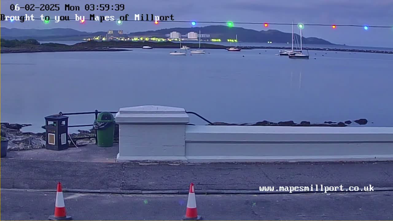 A calm, twilight view of a harbor with several boats, a low white wall, and two traffic cones in the foreground.