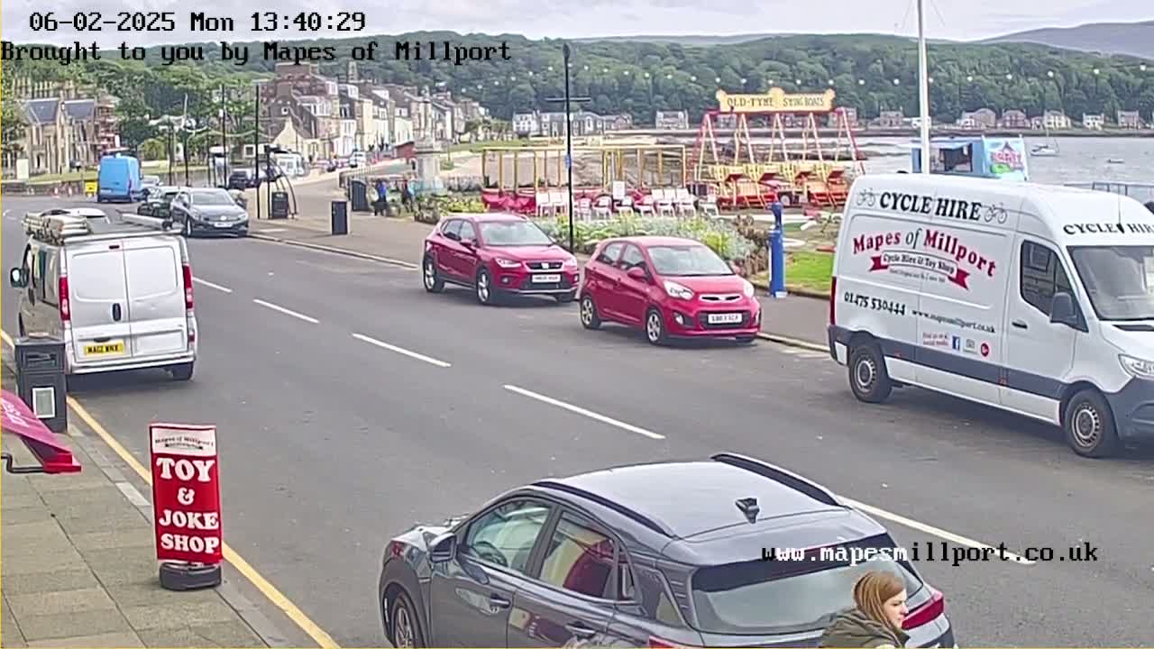 A partly cloudy day shows several cars parked and driving on a street near a seaside town with a carousel in the background.