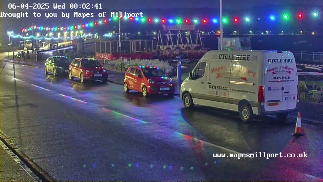 At night, under wet conditions, three cars and a white van are parked on a street illuminated by colorful lights strung overhead.