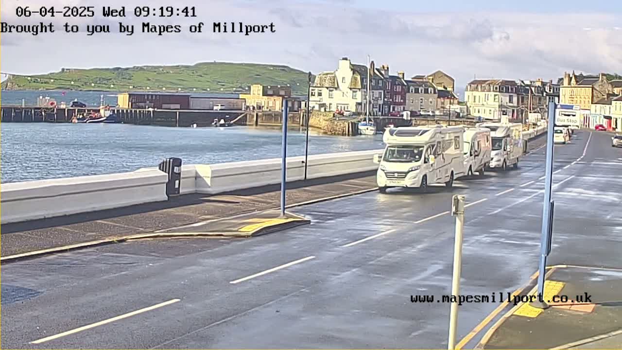 Several motorhomes are parked along a seaside road next to a harbor, under a partly cloudy sky.